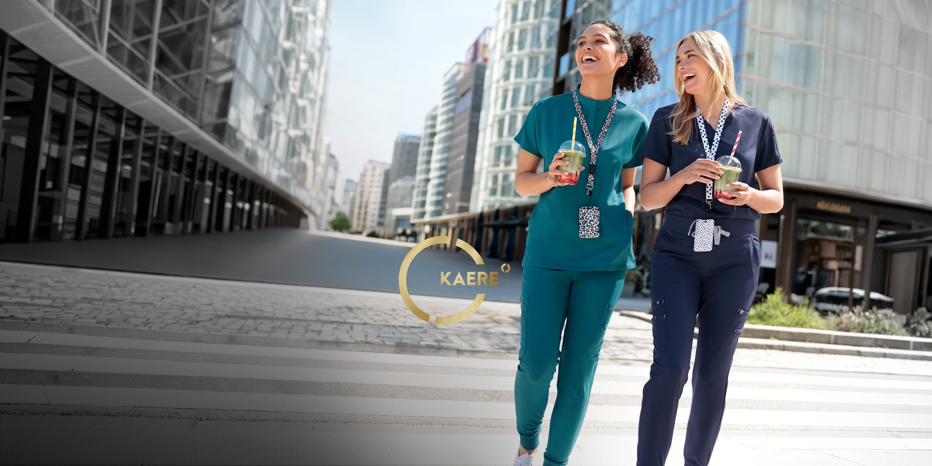Zwei Frauen in türkisfarbener und dunkelblauer KAERE Berufsbekleidung mit Getränken in moderner Stadtkulisse.